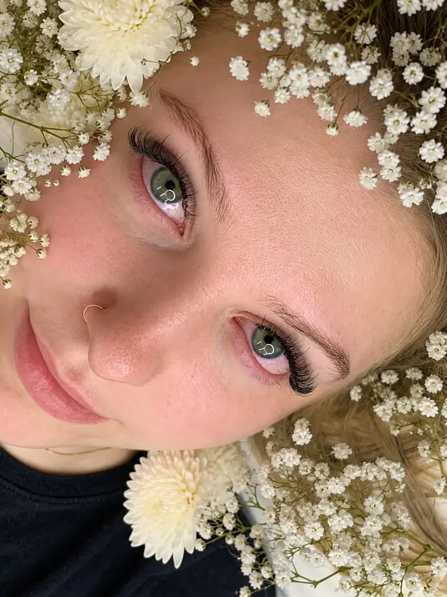 Cliente com extensão de cílios e sobrancelha desenhada, entre flores brancas